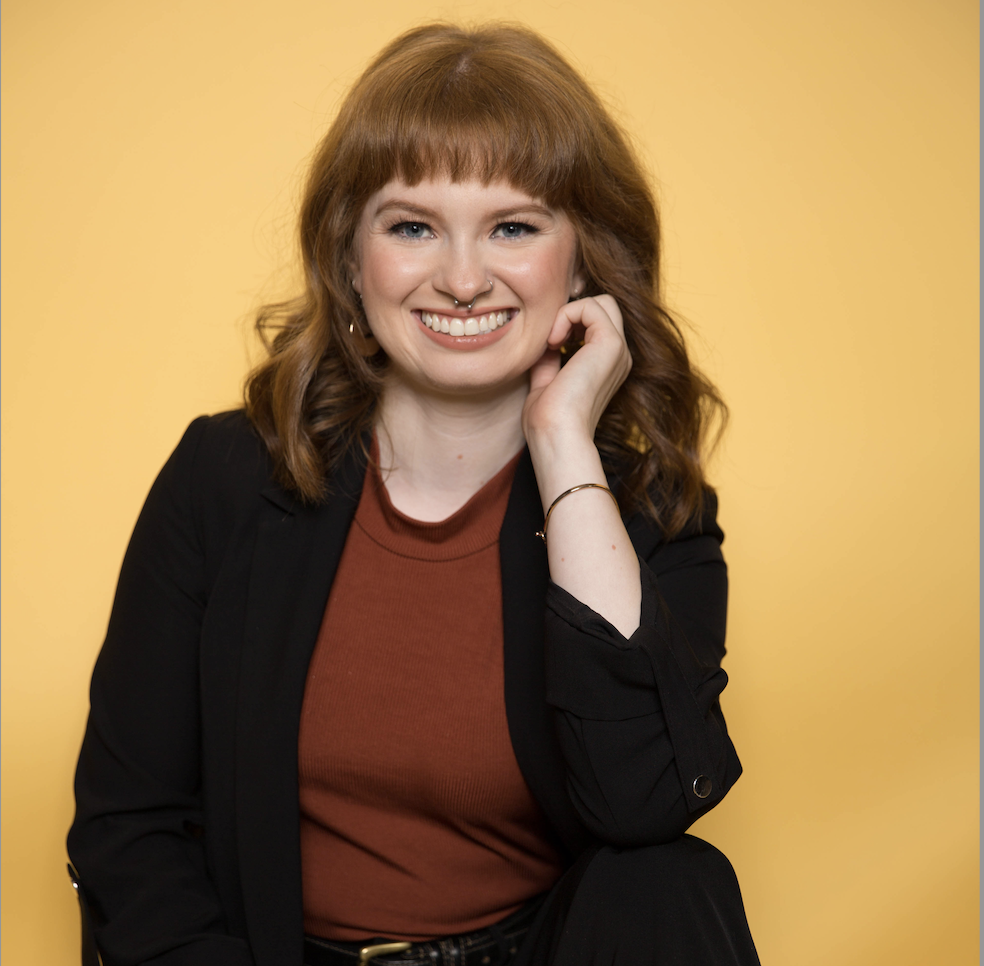 Smiling red-haired woman with bangs in black blazer and rust top, hand on cheek, yellow backdrop